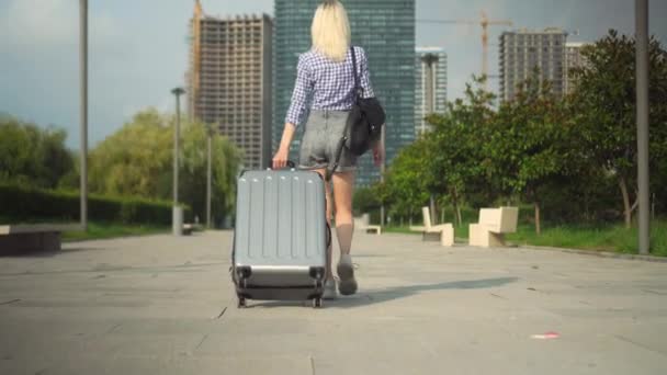 молодая красивая женщина проходит через курортный город и катит чемодан позади нее. - Кадры, видео