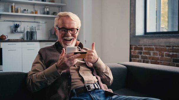 boldog nyugdíjas hitelkártyával mobiltelefon, miközben mutatja hüvelykujját fel otthon - Fotó, kép