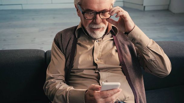 Emekli adam gözlüklerini ayarlıyor ve akıllı telefona bakıyor.  - Fotoğraf, Görsel