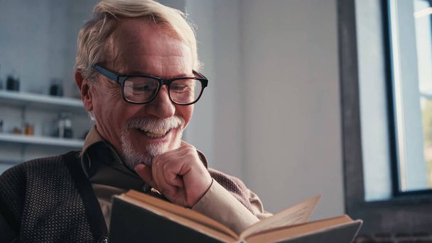 Χαρούμενος ηλικιωμένος άνδρας με γυαλιά ηλίου που διαβάζει βιβλία στο σπίτι  - Φωτογραφία, εικόνα