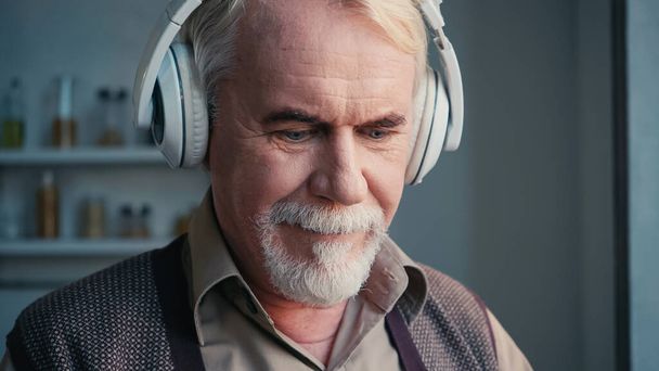 hombre barbudo y mayor en auriculares inalámbricos escuchando música y mirando hacia abajo - Foto, Imagen