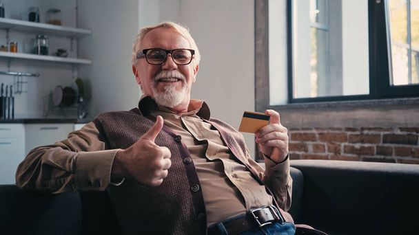 boldog nyugdíjas szemüvegben, aki hitelkártyát tart és hüvelykujját felfelé mutatja - Fotó, kép