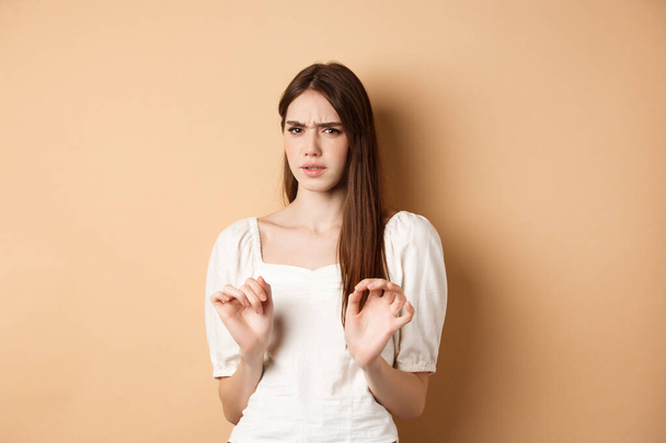 Stay away from me. Disgusted young woman frowning and looking with aversion at something stinky, rejecting disgusting thing, standing on beige background - Foto, imagen