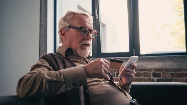 пенсіонер в окулярах використовує смартфон і тримає кредитну картку вдома
 - Фото, зображення