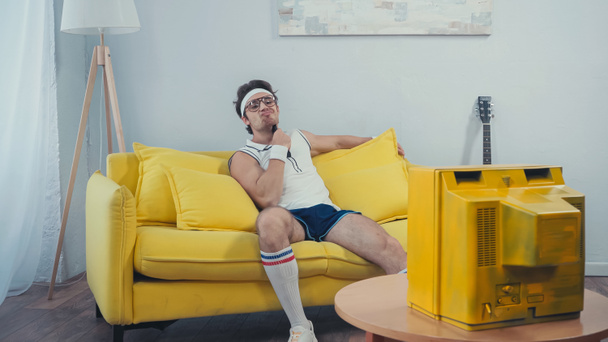 luie man in sportkleding krassen nek met afstandsbediening tijdens het zitten op de bank in de woonkamer - Foto, afbeelding