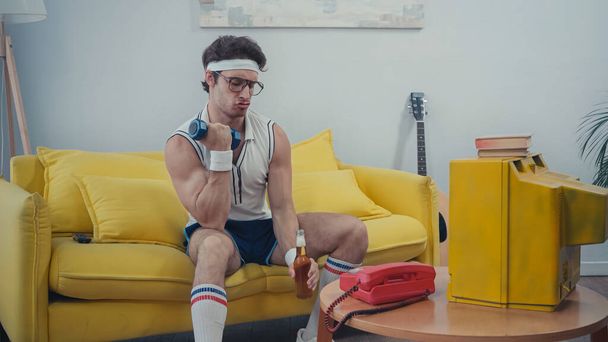 sportive man drinking beer and lifting dumbbell while sitting on sofa - Φωτογραφία, εικόνα