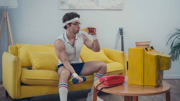 deportista beber cerveza y hacer ejercicio con la mancuerna mientras mira la televisión - Foto, imagen