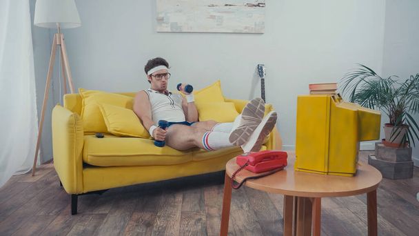 man met bril training met halters terwijl hij op de bank zit en tv kijkt in de woonkamer - Foto, afbeelding