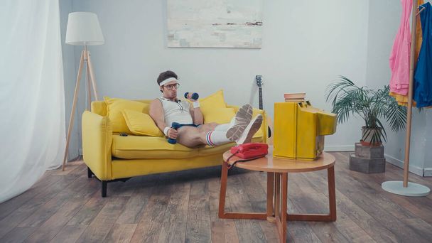 man met bril die met halters traint terwijl hij op de bank zit en tv kijkt in de woonkamer - Foto, afbeelding