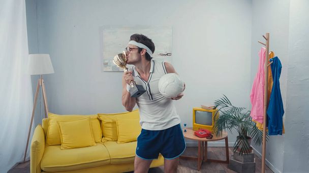 Αθλητής φιλιά κύπελλο τρόπαιο και σπορ μπάλα, ενώ στέκεται στο σπίτι - Φωτογραφία, εικόνα
