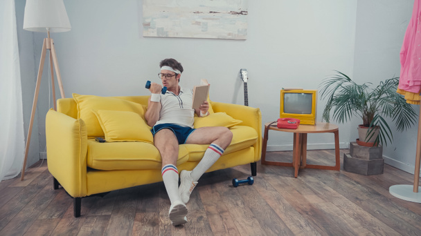 Esportista exercitando com haltere enquanto segurando livro na sala de estar - Foto, Imagem