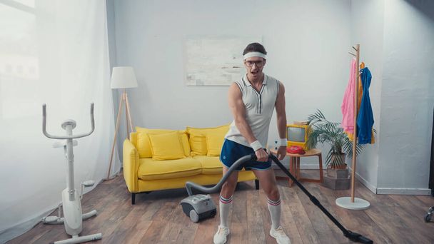 Sportsman singing and dancing while vacuum cleaning living room, retro sport concept - Photo, Image