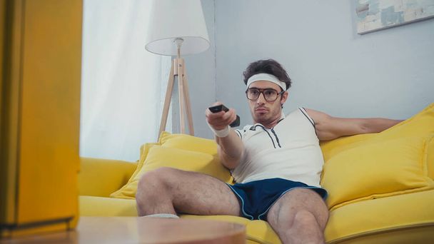 скучающий мужчина в ретро спортивной одежде сидит на диване и переключает каналы  - Фото, изображение