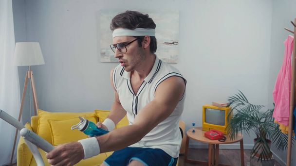 sportsman working out on exercise bike and holding sports bottle in living room, retro sport concept - Foto, afbeelding