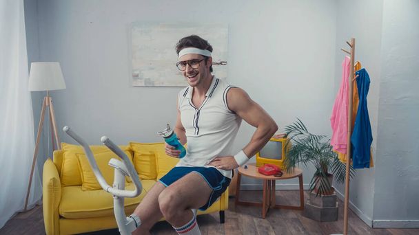 entrenamiento de deportista feliz en bicicleta estática y celebración de botella deportiva en la sala de estar, concepto de deporte retro - Foto, Imagen