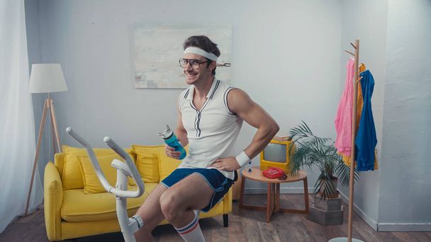 cheerful sportsman training on exercise bike and holding sports bottle in living room, retro sport concept - Photo, Image