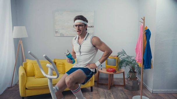 sportsman training on exercise bike and holding sports bottle in living room, retro sport concept - Photo, Image