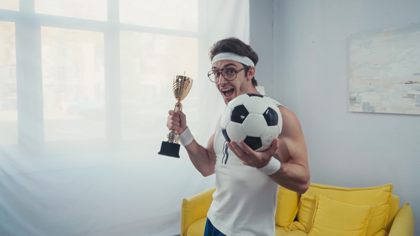 Excited football player with trophy cup and soccer ball celebrating win at home - Photo, Image
