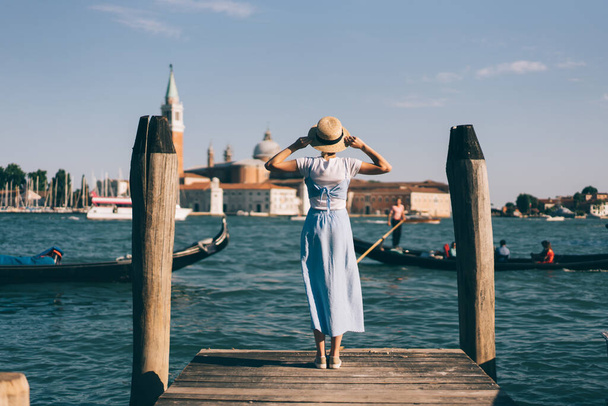 Back view of woman in dress touching hat while standing on wooden pier near broad channel with gondolas in Venice, Italy - Foto, Imagem