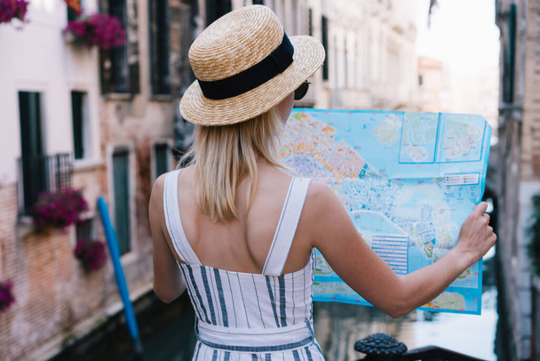Back view of young blond female tourist in hat looking map and checking location while standing in Venice street in Italy - Photo, image