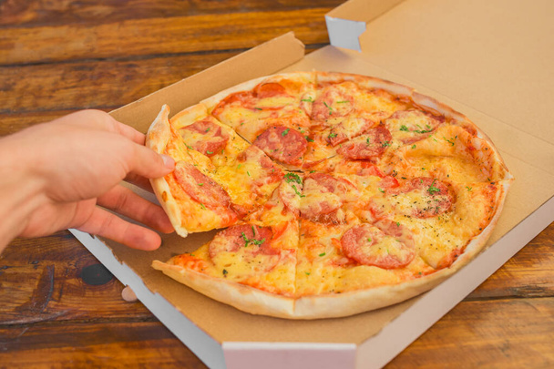 Рука парня берет пиццу в картонной коробке на фоне деревянного стола. Вкусный фаст-фуд. - Фото, изображение