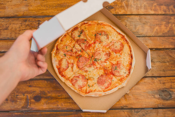 La mano del ragazzo apre una scatola di cartone di pizza sullo sfondo di un tavolo di legno. Delizioso fast food. - Foto, immagini