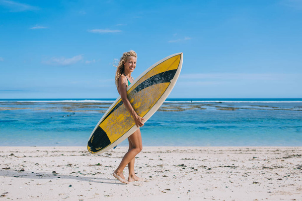 Side view of cheerful youthful sporty light haired woman looking at camera and holding surf board while standing during vacation on coastline - Fotó, kép