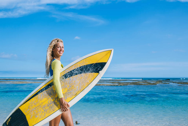 Side view of beautiful tanned female with long blonde hair holding yellow and black surfboard while standing on tropical beach and enjoying summer holiday - Zdjęcie, obraz
