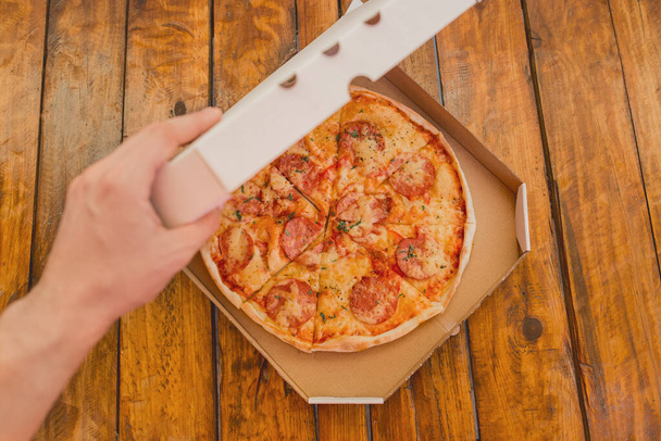 Ręka faceta otwiera kartonowe pudełko pizzy na tle drewnianego stołu. Pyszne fast food. - Zdjęcie, obraz