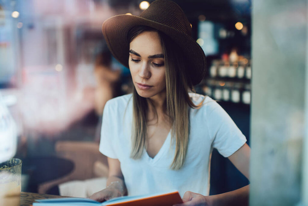 Concentrated and smart woman in T-shirt and trendy hat sitting in cafe and attentively reading book while enjoying new interesting story  - Φωτογραφία, εικόνα