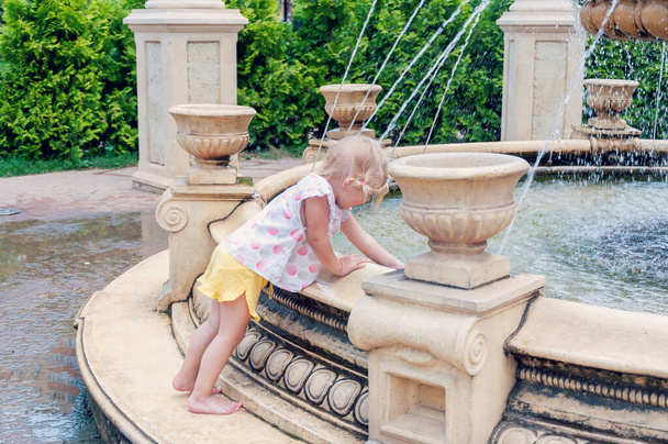 Near The Fountain. Girl having fun with fountain at hot sunny day. Cute little girl playing with fountain splash - Photo, image