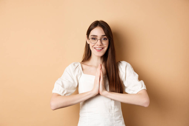 Smiling girl in glasses say thank you, show namaste gesture, express gratitude, standing pleased on beige background - Photo, Image