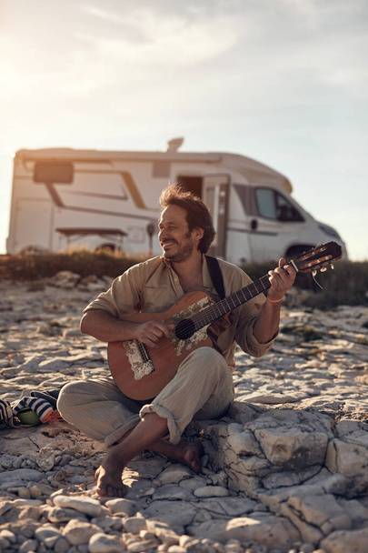夏には海の海岸でギターを演奏しているキャンパーハウスを持つヒッピーギタリスト. - 写真・画像