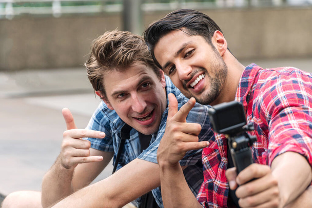 Dois blogueiros amigos bonitos tirando selfie junto com a câmera de ação sentada na cidade de rua olhando a câmera fazendo cara de prazer. Dois viajantes felizes tiram fotos enquanto viajam de férias. selecionar foco - Foto, Imagem