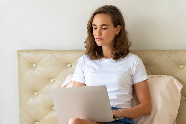 Concepto tecnológico. Blogger joven mujer que trabaja en línea con el ordenador portátil mientras está sentado en la cama en casa. Encantadora hembra usando notebook conectar internet buscando algo en la habitación. - Foto, imagen