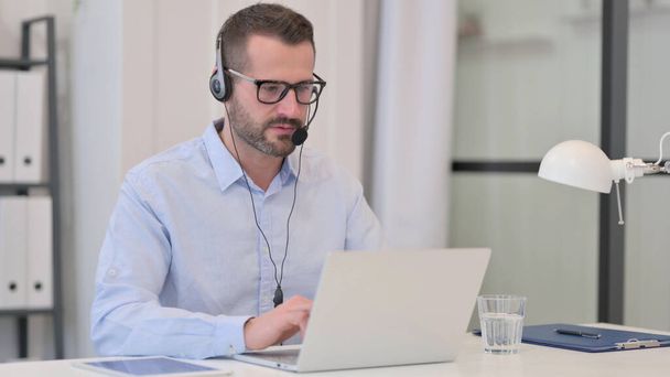 Człowiek w średnim wieku z słuchawkami Rozmowa podczas pracy na laptopie  - Zdjęcie, obraz