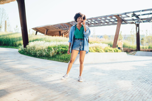 Happy carefree young black lady in shorts and shirt standing in tranquil place in park and chatting on smartphone looking at camera - Fotografie, Obrázek