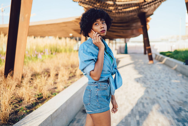 Pretty dark skinned woman with curly hair talking on mobile phone walking outdoors during summer day, trendy dressed african american hipster girl traveler making call in roaming during trip - Foto, immagini