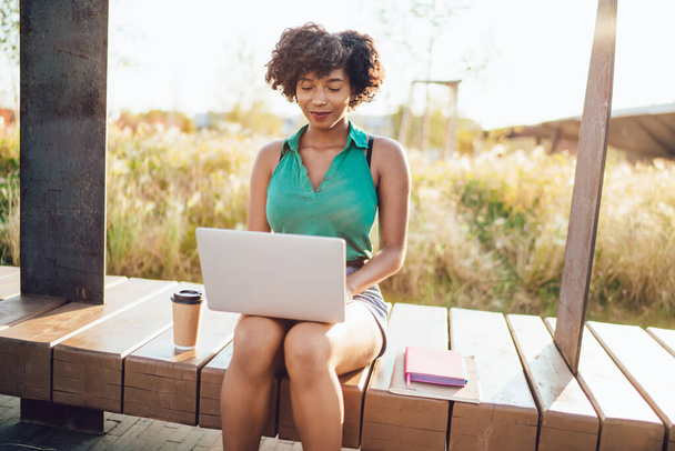 Prospera mujer de piel oscura freelancer escribiendo en el ordenador portátil pasar el día soleado en el uso del parque Internet 4G, mujer afroamericana positiva viendo webinar aprendizaje utilizando la tecnología en el ocio - Foto, imagen