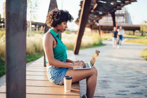 Focused ethnic lady with curly hair dressed in green shirt and shorts surfing laptop sitting with cup of coffee in street in summer  - Photo, image