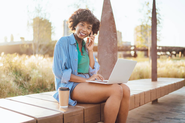 Portrait of cheerful african american woman freelancer talking on mobile phone about business online sitting in park, charming dark skinned female blogger satisfied with 4G internet use gadgets - Photo, image