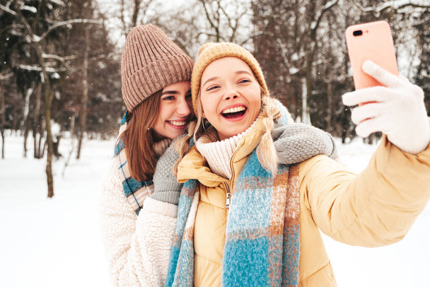 Two young beautiful smiling hipster female in trendy warm clothes and scarfs.Carefree women posing in the street in park. Positive pure models having fun in snow. Enjoying winter moments.Taking selfie - Фото, изображение