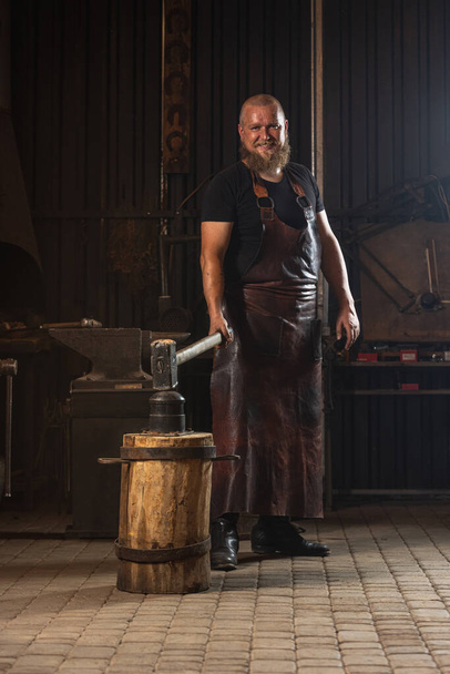 Full ength portrait of bearded man, blacksmith working on creating handmade metal product at family smithy. Concept of labor, retro professions, power, beauty - 写真・画像