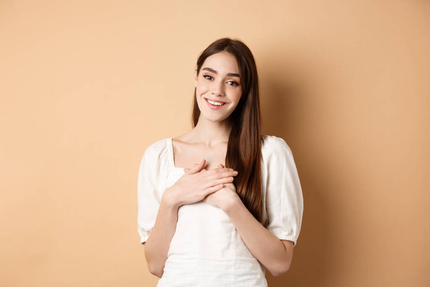 Pretty girl holding hands on heart and smiling, thanking you, feeling grateful and pleased, standing in dress on beige background - Foto, Imagen