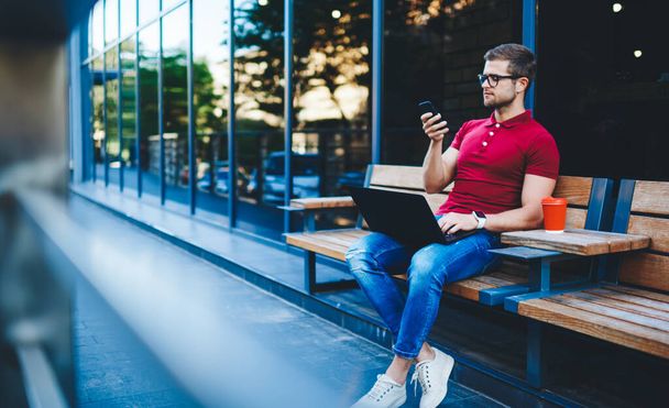 Trendy dressed caucasian male freelancer checking income message on smartphone working remotely outdoors, pensive millennial hipster guy student sitting with digital devices on street use 4G internet - Фото, зображення