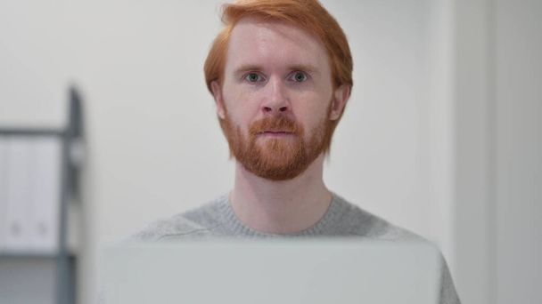 Бородатый рыжий с ноутбуком смотрит в камеру  - Фото, изображение