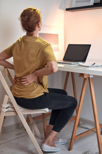 Kobieta pracująca na laptopie w domu i mająca problemy z biodrami, bólem pleców i brzucha. - Zdjęcie, obraz