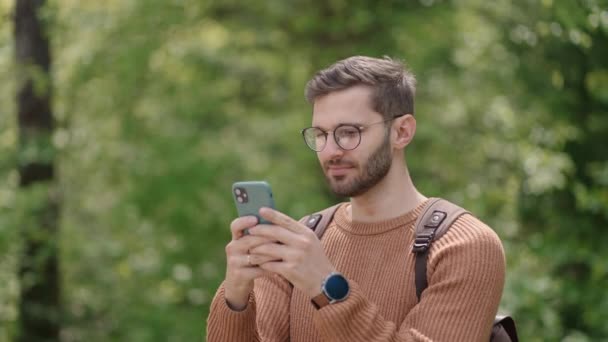 Mladý muž s batohem v lese, v přírodě píše vzkazy na mobilní telefon. Turista na otevřeném prostranství komunikuje v chatu, dívá se na fotografie, raduje se, usmívá. - Záběry, video