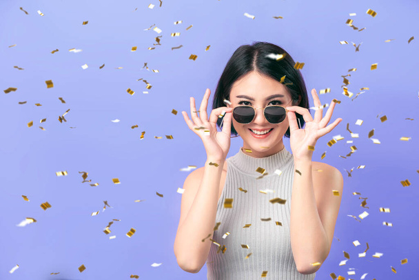 Щаслива жінка з Азії у сонцезахисних окулярах посміхається з святковим сюрпризом конфетті на фіолетовому фоні. - Фото, зображення