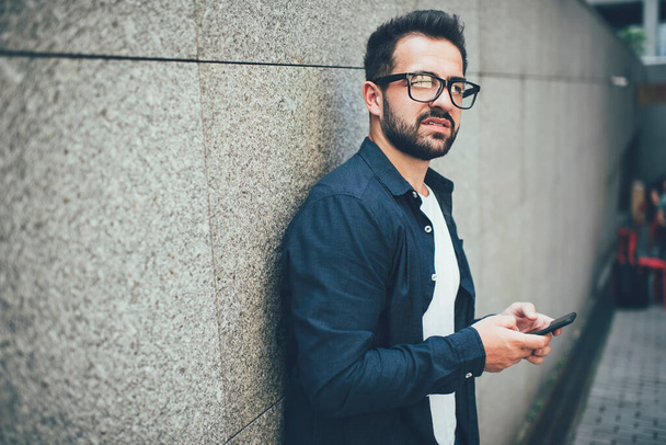 Pensive caucasian hipster guy recalling password for enter app banking account on mobile phone standing outdoors, pondering millennial man in casual wear forgot number for dialing on cellular - Fotografie, Obrázek